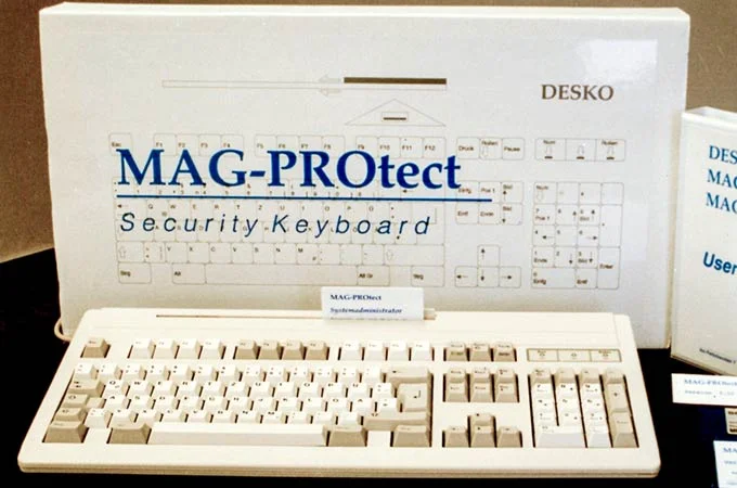 Produktfoto einer beigen DESKO &bdquo;MAG-PROtect&ldquo; Security-Tastatur mit Verpackung und Benutzerhandbuch im Hintergrund.