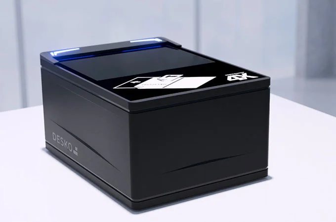 Ein kompakter schwarzer DESKO-Dokumentenscanner steht auf einem Tisch, die beleuchtete Scanfl&auml;che ist oben sichtbar.