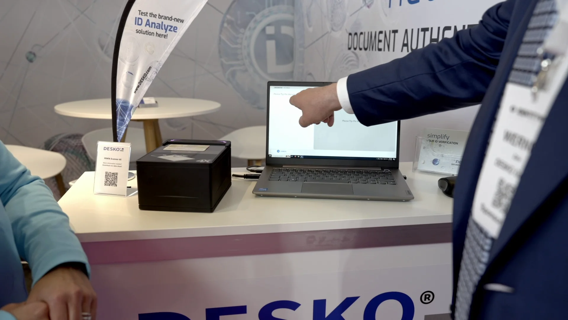 Man points to laptop with ID data, next to document scanner at the stand for identity verification.