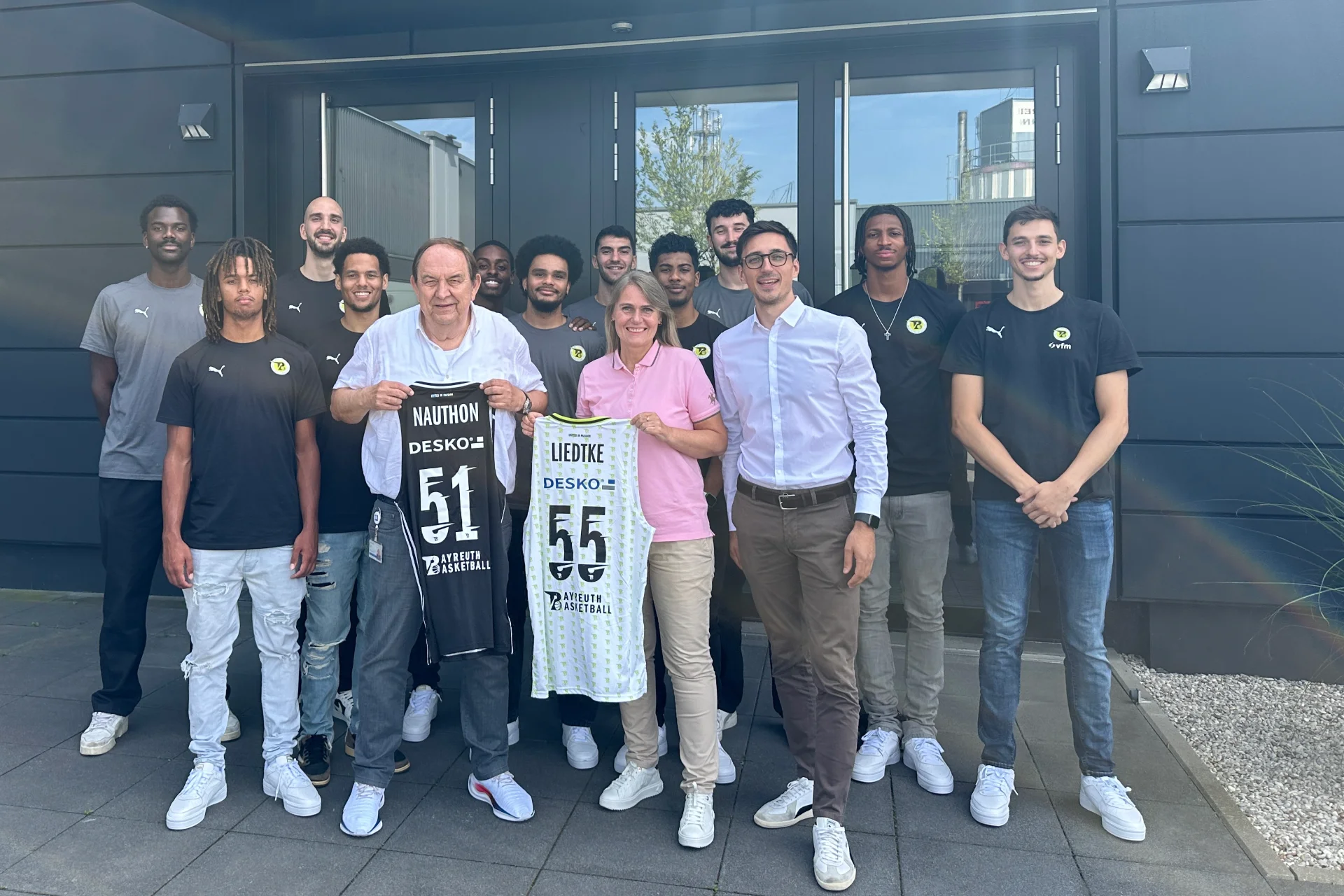 Gruppe von Basketballspielern und Partnern steht vor einem Geb&auml;ude und pr&auml;sentiert zwei Bayreuth-Basketball-Trikots.