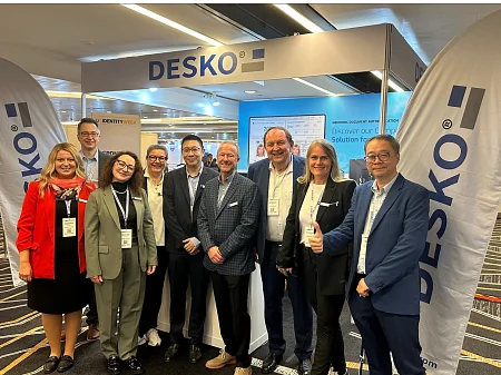Gruppe von DESKO-Mitarbeitern und Partnern steht lächelnd vor einem DESKO-Messestand mit Bannern und Präsentationsscreen.