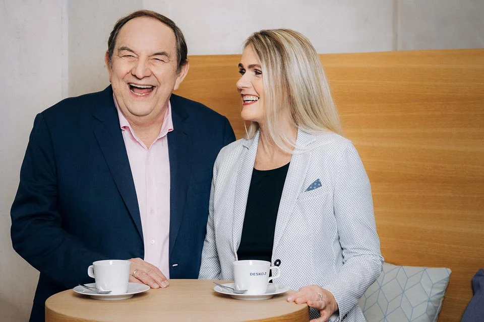 Zwei Personen sitzen lachend an einem Tisch, trinken Kaffee aus Tassen und unterhalten sich in entspannter Atmosph&auml;re.