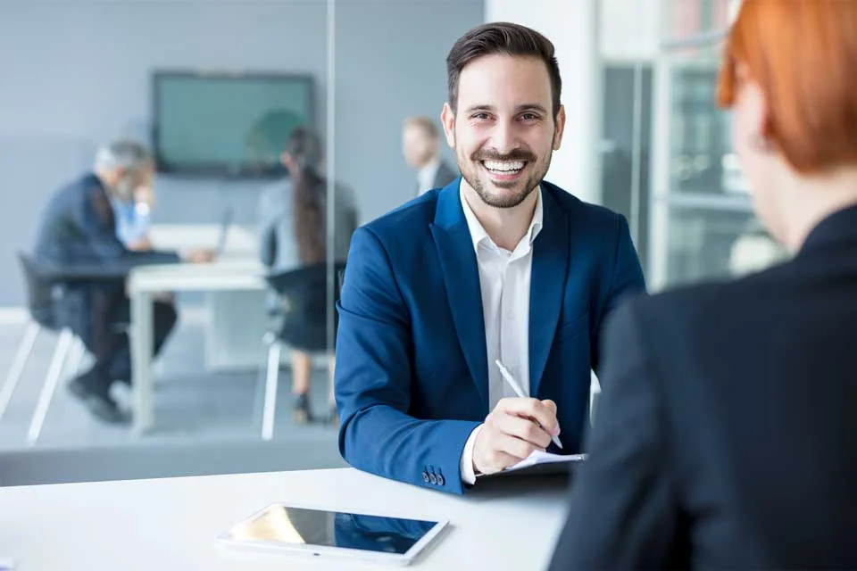 Mann im Anzug sitzt an einem Tisch und f&uuml;hrt ein Beratungsgespr&auml;ch mit einer Person in einem modernen B&uuml;ro.
