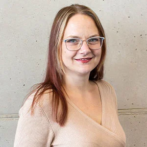Portr&auml;t einer l&auml;chelnden Frau mit Brille und langen Haaren vor hellem Betonhintergrund.