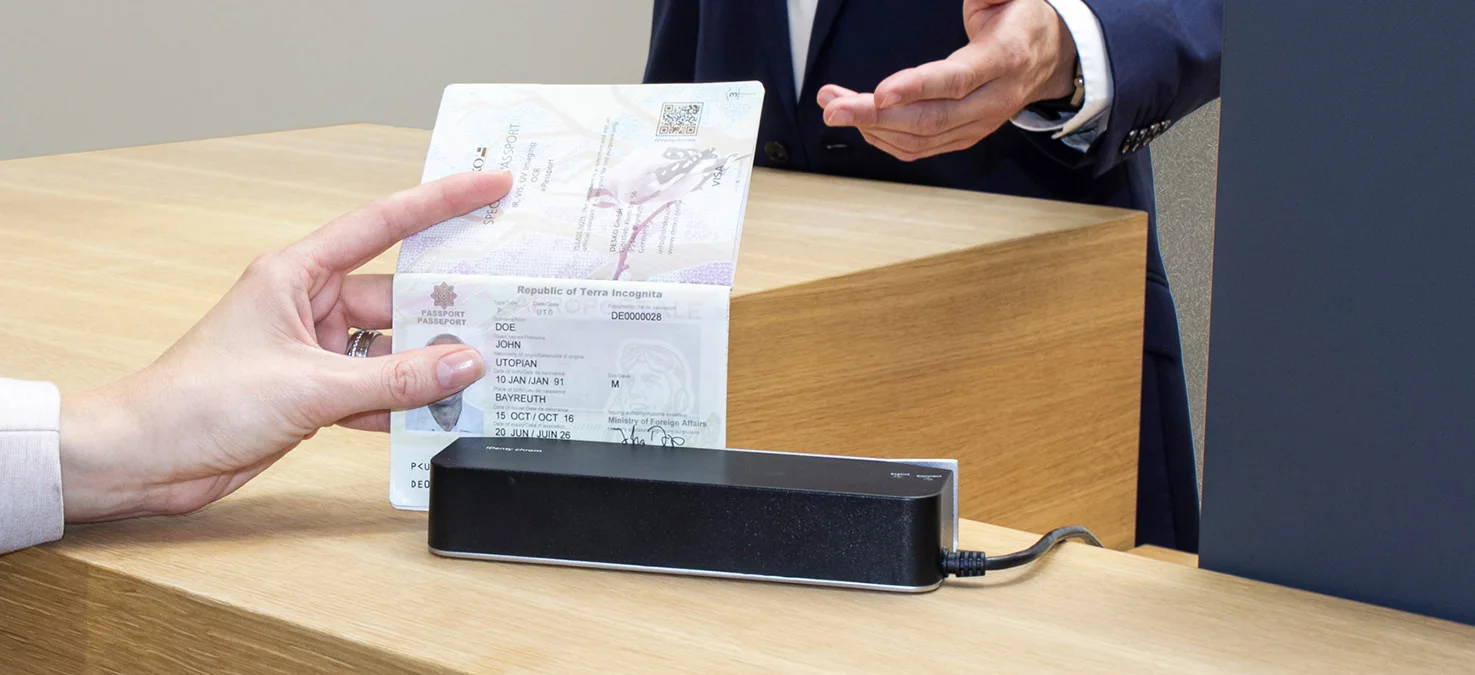 Main tenant un passeport ouvert au-dessus d'un lecteur au guichet, tandis qu'un employ&eacute; derri&egrave;re lui demande de v&eacute;rifier l'identit&eacute;.