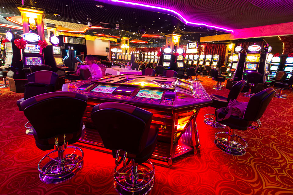 Raum mit Spielautomaten an den Wänden und in der Mitte ein Roulette-Tisch mit Stühlen
