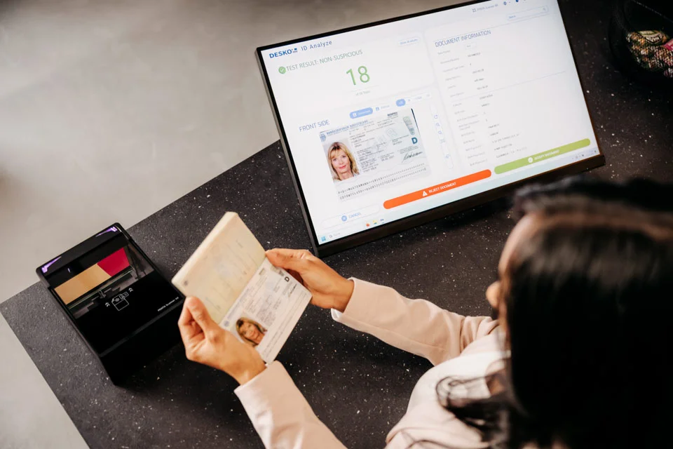 Frau mit Reisepass in der Hand steht vor einem Passscanner und einem Monitor der eine Pr&uuml;fsoftware zeigt - DESKO ID Analye