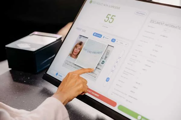 Person zeigt mit dem Finger auf eine digitale Ausweisanalyse auf einem Bildschirm, daneben steht ein Dokumentenleseger&auml;t auf dem Tresen.