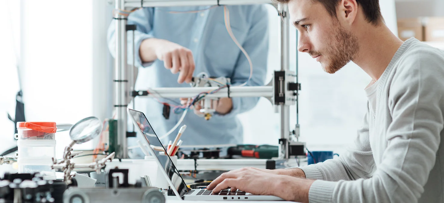 Un joven se concentra en su port&aacute;til mientras un colega trabaja en un prototipo t&eacute;cnico en segundo plano.