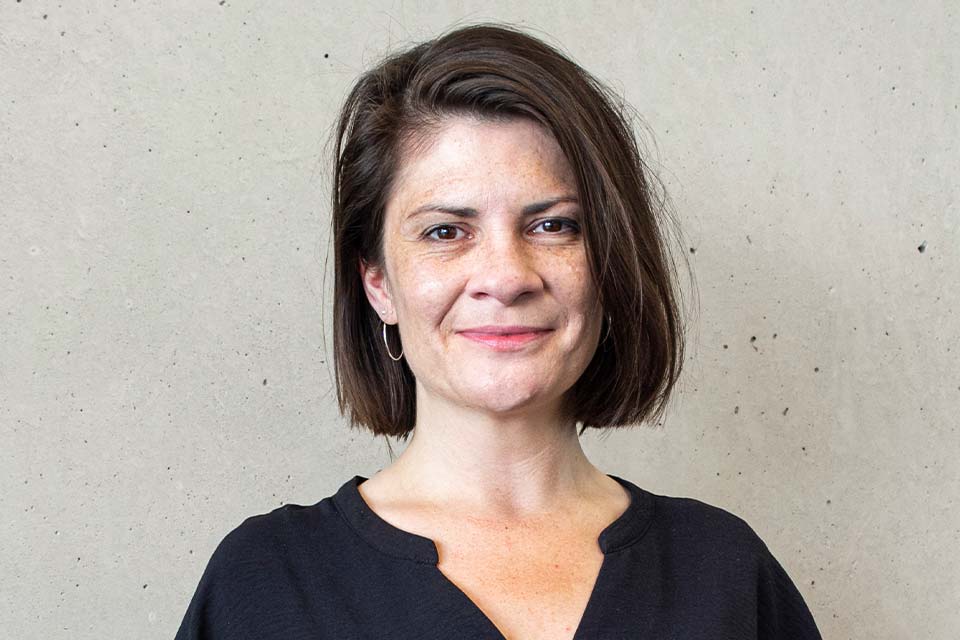 Porträt einer lächelnden Frau mit blonden Haaren und runder Brille vor unscharfem, modernem Bürohintergrund.