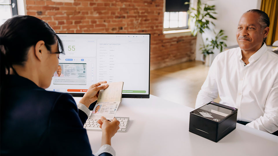 A woman is looking at the passport of a customer sitting opposite her. A passport scanner is on the table in front of her, and on the screen next to her, you can see verification software.