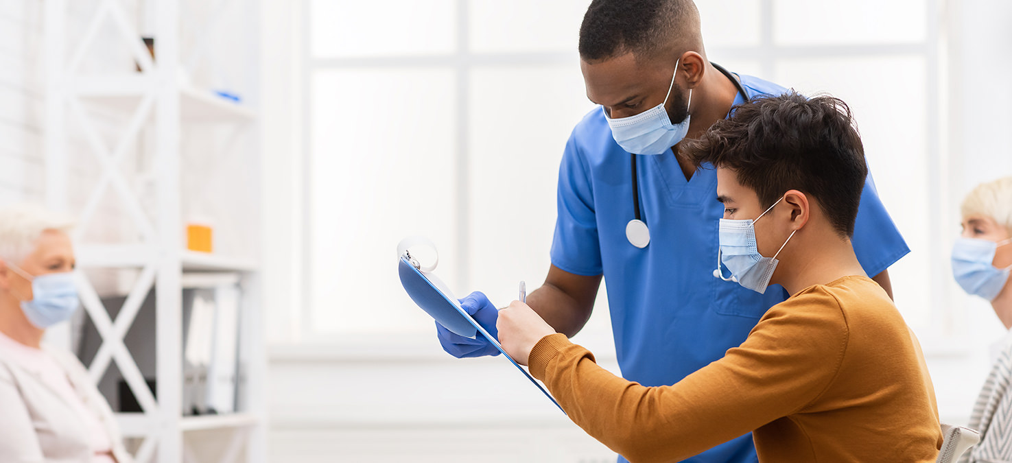 Patienten und Pfleger unterschiedlicher Ethnien in Wartezimmer. Alle tragen Mund-Nasen-Masken.