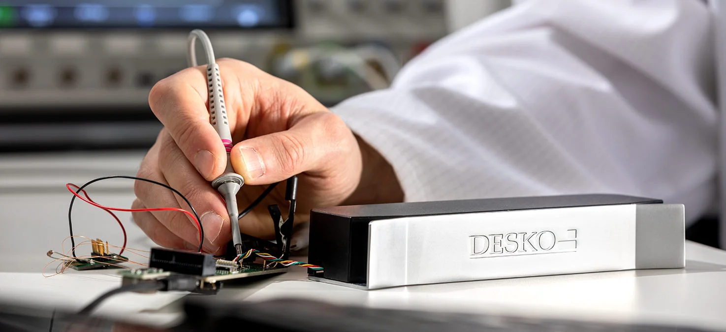 El t&eacute;cnico mide con puntas de prueba en una placa electr&oacute;nica abierta, un dispositivo DESKO descansa sobre una superficie de trabajo junto a ella.