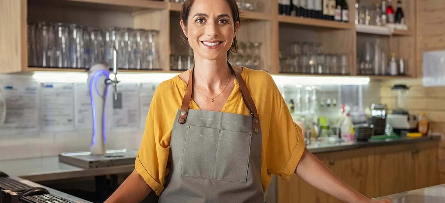 Freundliche Servicekraft mit Sch&uuml;rze steht hinter einem Tresen in einem Caf&eacute; oder Bistro, im Hintergrund Gl&auml;ser und Getr&auml;nkeflaschen.