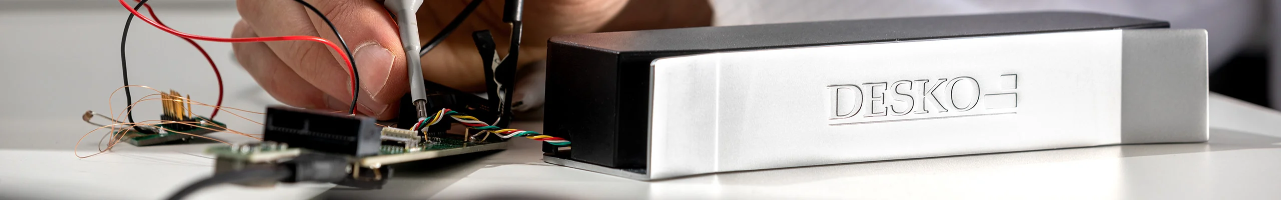Un technicien prend des mesures avec des pointes de test sur une carte &eacute;lectronique ouverte, &agrave; c&ocirc;t&eacute; d'un appareil DESKO pos&eacute; sur un plan de travail.