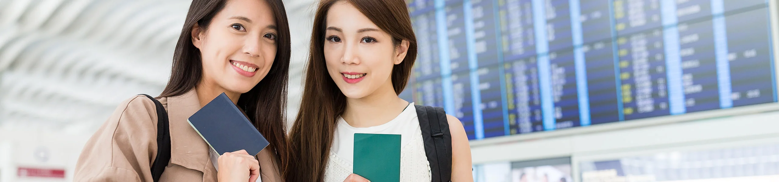 Two women with rucksacks hold passports in their hands and stand in front of a large display board showing departure times at the airport.
