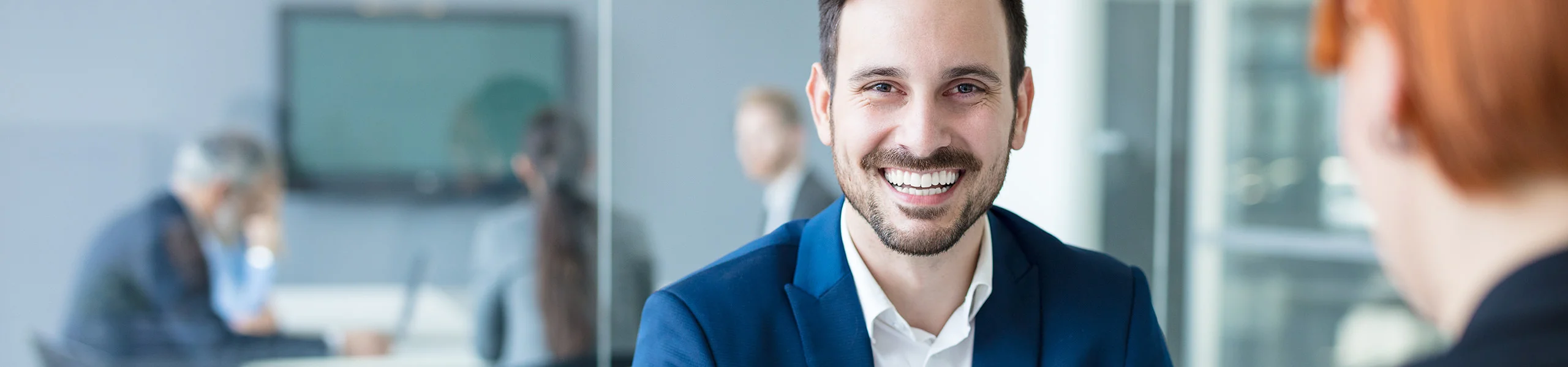 Hombre sonriente con traje sentado en una sala de reuniones hablando con un colega, otras personas difuminadas en el fondo.