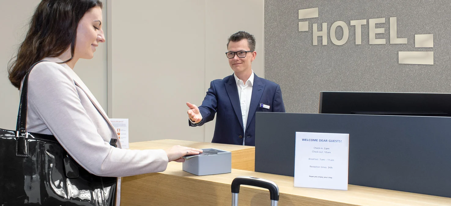 Hotelgast legt eine Karte auf ein kontaktloses Leseger&auml;t am Empfangstresen, w&auml;hrend ein Mitarbeiter hinter dem Tresen assistiert.