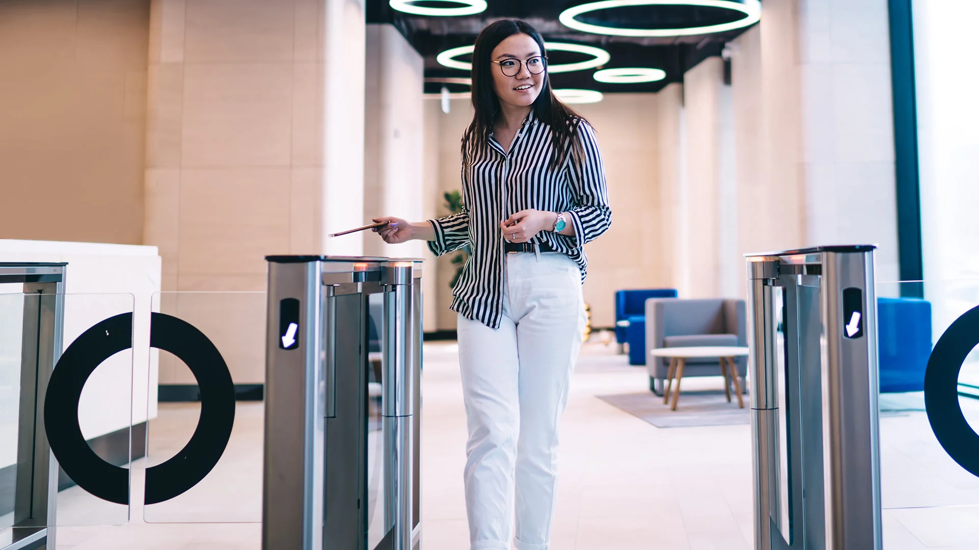 Mujer con gafas pasa por un torniquete electr&oacute;nico con tarjeta de acceso en una oficina moderna con l&aacute;mparas redondas en el techo.