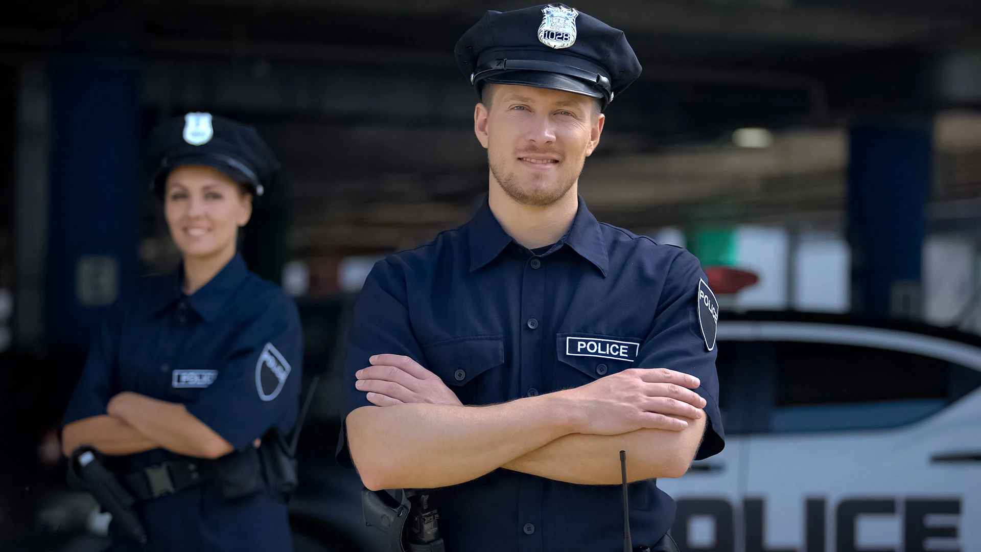 Un policier et une polici&egrave;re se tiennent debout, les bras crois&eacute;s, devant une voiture de police.