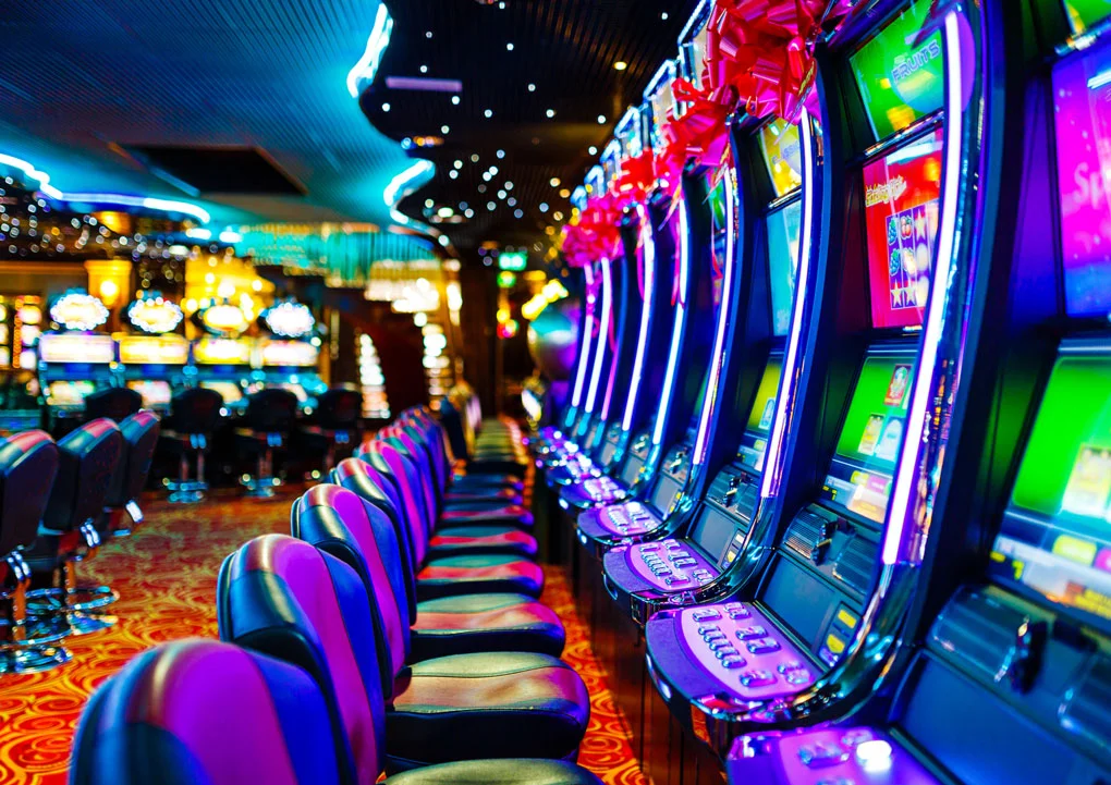 Reihe moderner Spielautomaten in einem Casino, bunt beleuchtet mit Neonfarben, leere Sitze davor und glitzernde Spielatmosph&auml;re.