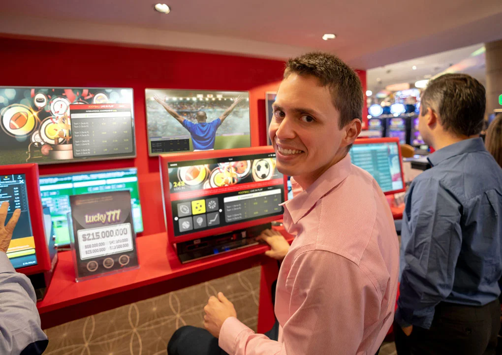 L&auml;chelnde Person sitzt vor roten Casino-Terminals mit Sportwetten- und Gl&uuml;cksspielanzeigen, weitere G&auml;ste spielen daneben.
