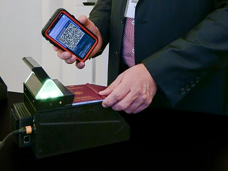Hand hält Smartphone mit QR-Code, während Reisepass auf beleuchtetem Ausweisscanner zur Prüfung liegt.