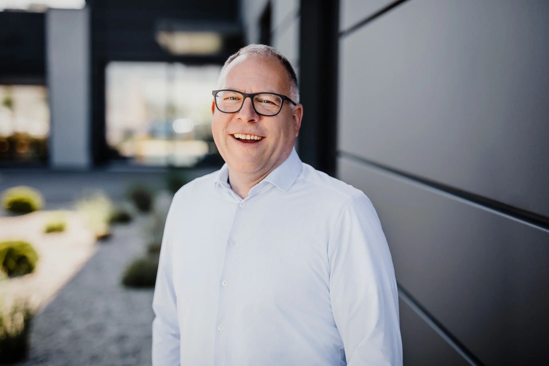 Smiling man with glasses and white shirt stands outside in front of a modern office building.