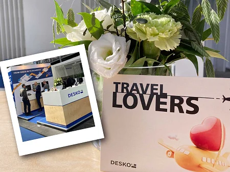 Blumenstrauß auf Tisch neben DESKO-Karte „Travel Lovers“, davor Polaroid-Foto eines DESKO-Messestands.