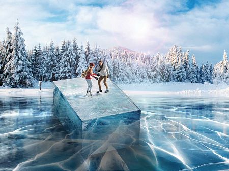 Two people glide on skates over a transparent block of ice on a frozen lake, surrounded by a snow-covered winter forest.